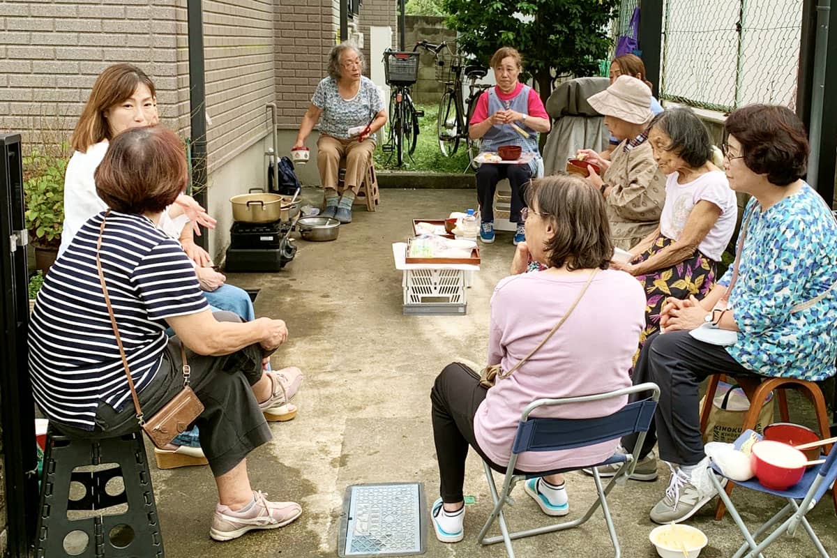 住居脇の駐車スペースにパイプ椅子などを並べて、あつまりを開いている。9人の女性たちが食事をしたり、会話をしている。