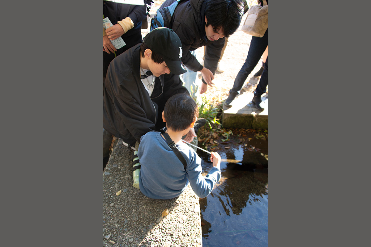 水路のところで子どもと高校生がしゃがんで、手を小さく開いて何かを観察している。