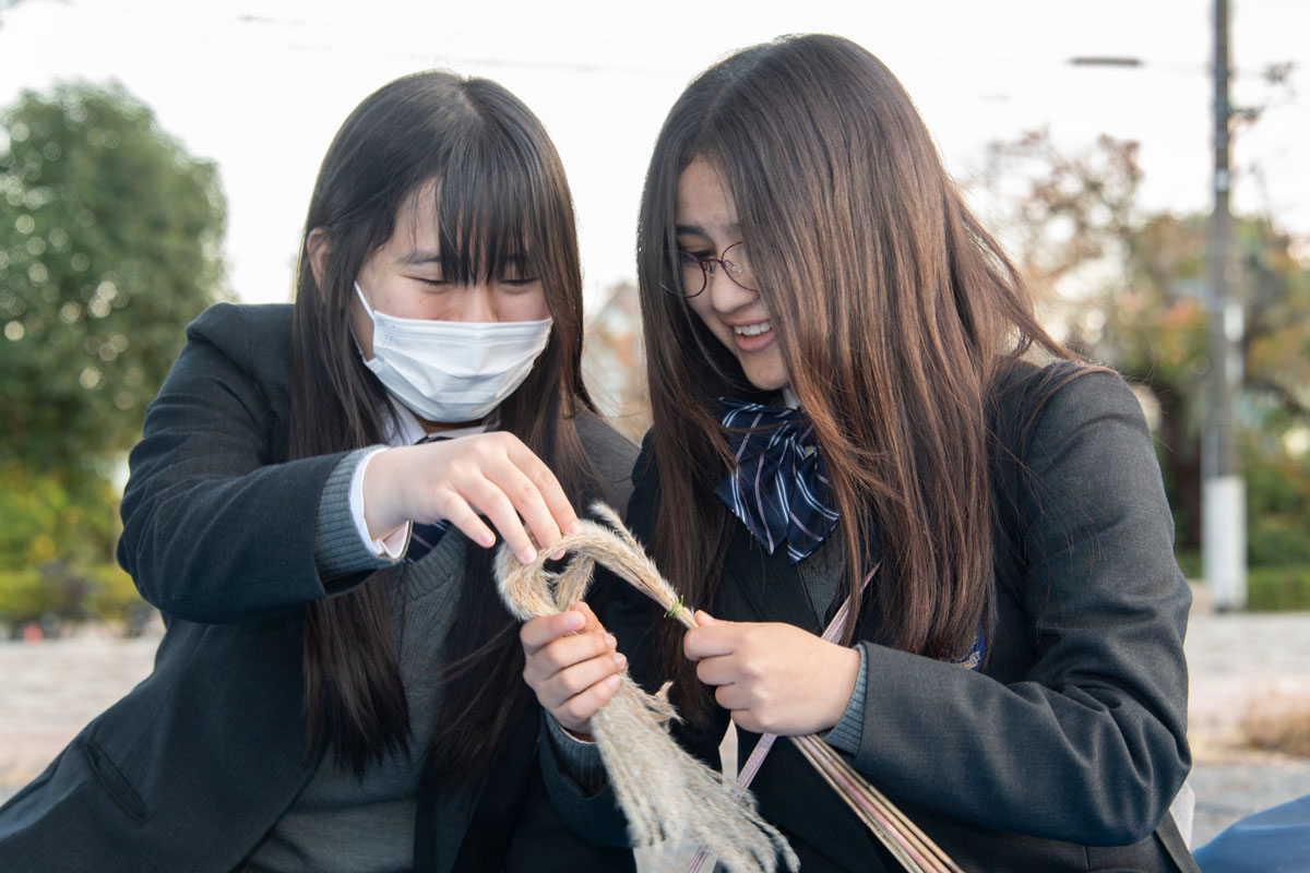 公園に広げたブルーシートの上で二人の高校生がにこやかな様子で、植物のオギを束ねて折り曲げている。