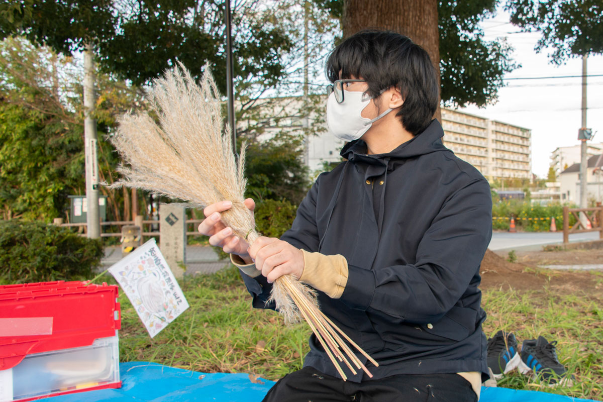 公園に広げたブルーシートの上で若い男性がススキのような見た目で、適度な長さにカットされたオギを束ねている。