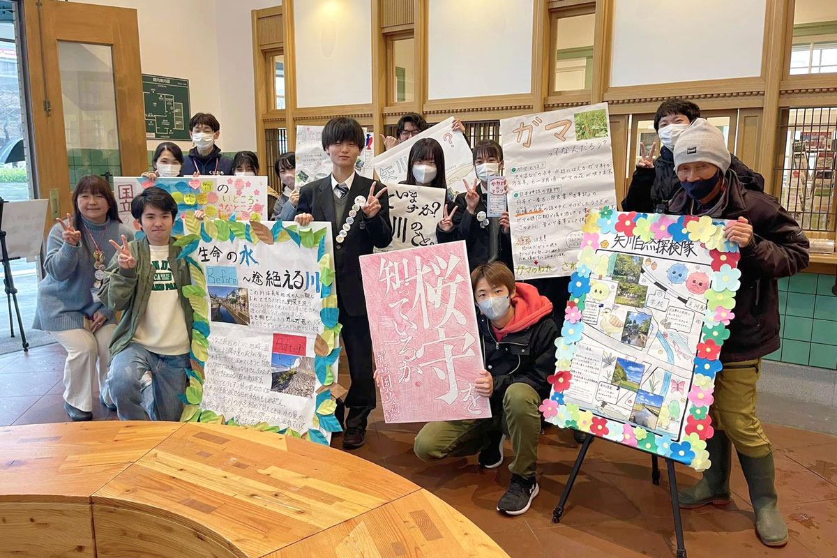 くにたち旧駅舎の建物内で、矢川自然探検隊のいろんな紹介や解説のポスターを持って高校生たちがみんなで写っている。大人もちらほら見える。